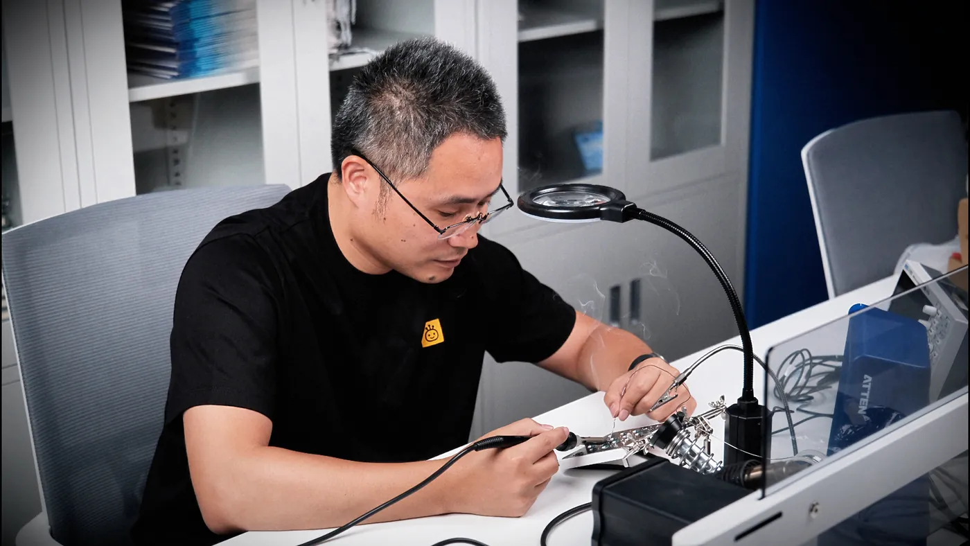 Tony at a workbench with a magnifier lamp and soldering iron, hand-soldering a board held in a helping-hands clamp